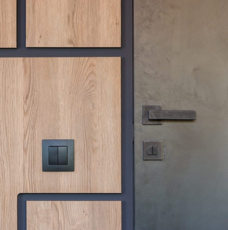 Close up image of a door and panel with earthy tones, featuring a black door handle and black light switch, showing simplicity in a modern style.
